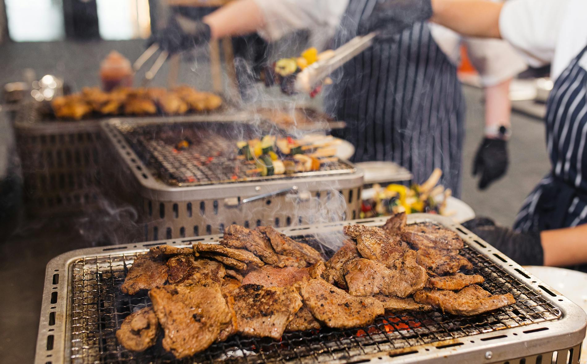 Te Pae chefs grill marinated meats on charcoal, filling the air with smoke. Behind them, colorful vegetable skewers sizzle on a second grill, creating a lively cooking environment.